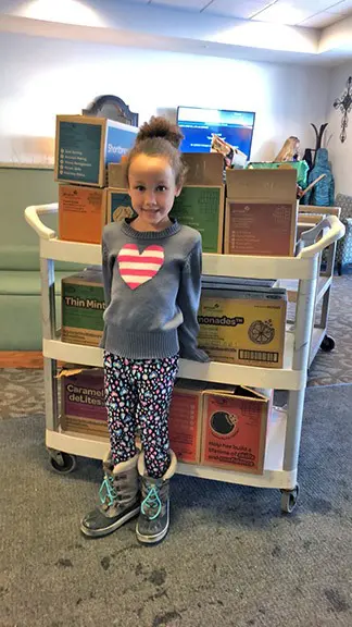 Decal Installer: A young Girl Scout stands in front of a cart stacked with boxes of cookies at the Good Samaritan Home in Waconia, with chairs and a TV in the background. – Image Graphics Premier Applicators Decal Installer: A young Girl Scout stands in front of a cart stacked with boxes of cookies at the Good Samaritan Home in Waconia, with chairs and a TV in the background. - Image Graphics Premier Applicators